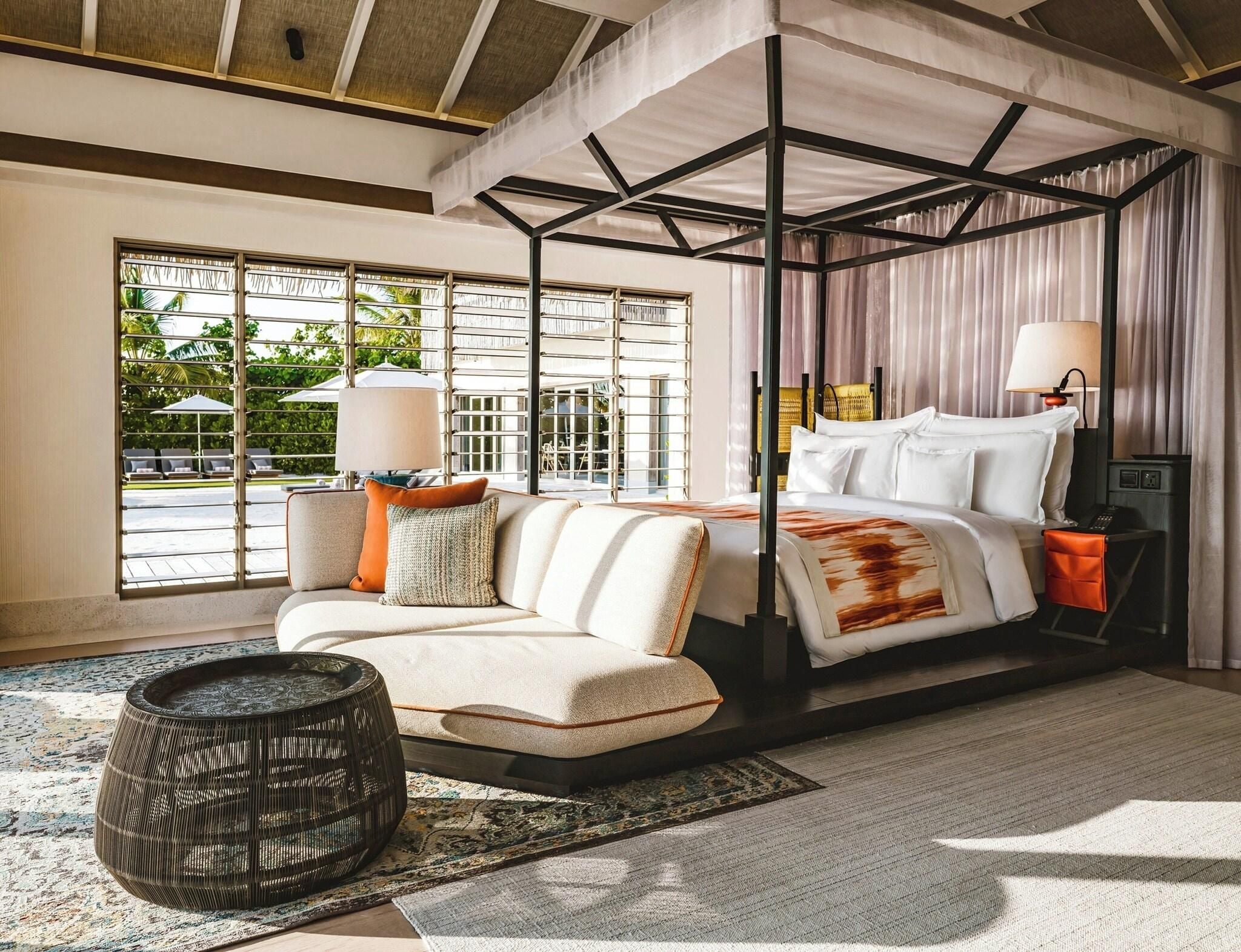 A four-poster bed with a canopy, a sofa with cushions, a side table with a lamp, and a view of the outdoors through large windows.