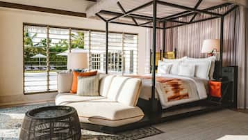 A four-poster bed with a canopy, a sofa with cushions, a side table with a lamp, and a view of the outdoors through large windows.