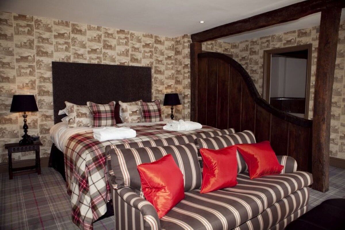 A bed with a headboard, a bedside table with a lamp, a bench with pillows, and a wooden door.