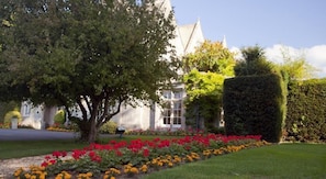 A building with a symmetrical facade, large windows, and a well-maintained garden with red and yellow flowers. - The Greenway (Cheltenham)