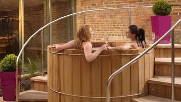 Two people in a wooden hot tub with metal railings, set against a brick wall and glass windows.