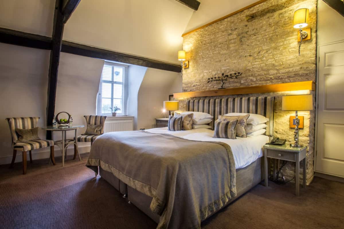 Un dormitorio con pared de piedra, una cama con cabecero, dos mesitas de noche con lámparas y una pequeña mesa con sillas.