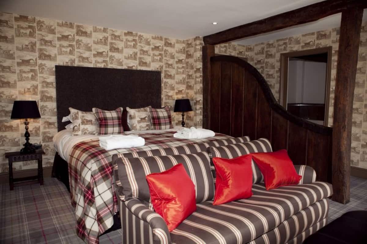 Una cama con cabecera, una mesita de noche con lámpara, un banco con almohadones y una puerta de madera.