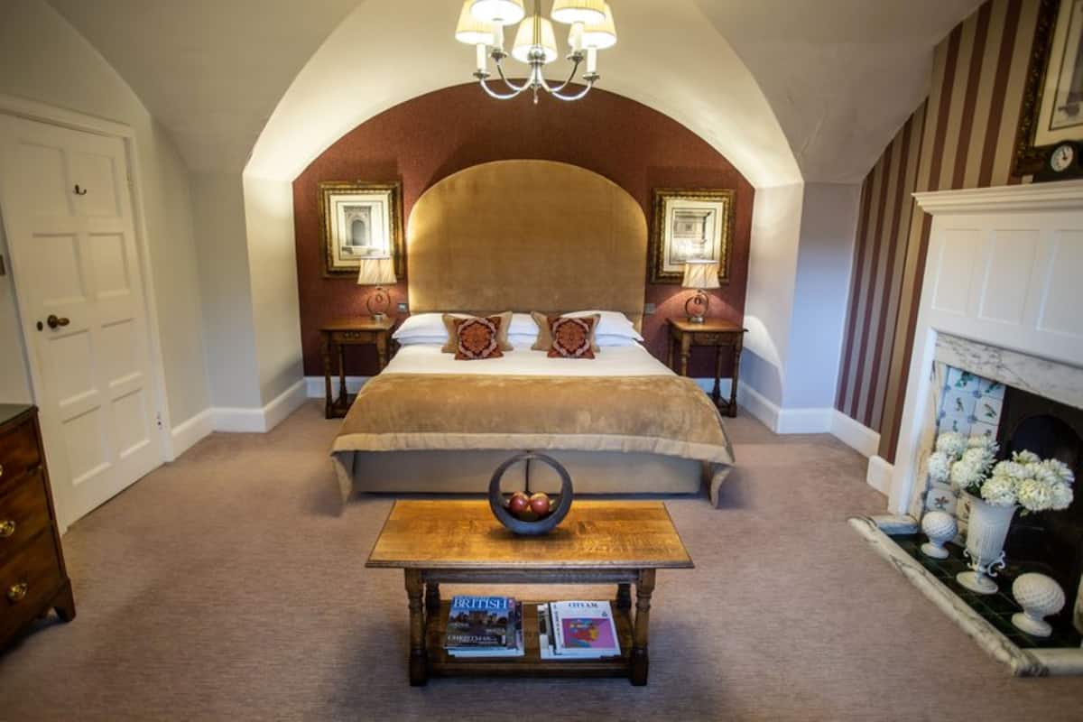 Un dormitorio con una cama grande, una araña, una chimenea y una pequeña mesa con libros.