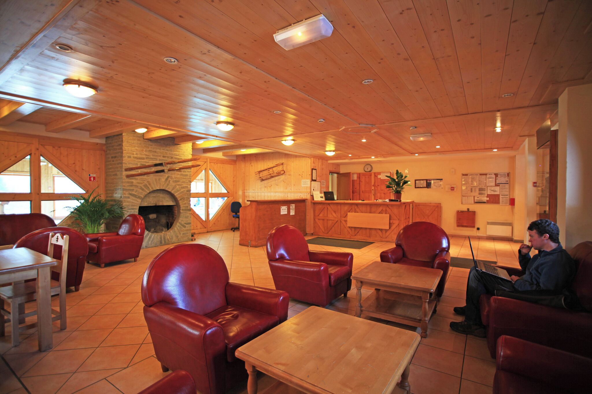 A cozy interior with wooden ceilings, red leather chairs, a fireplace, a reception desk, and a laptop on a table.