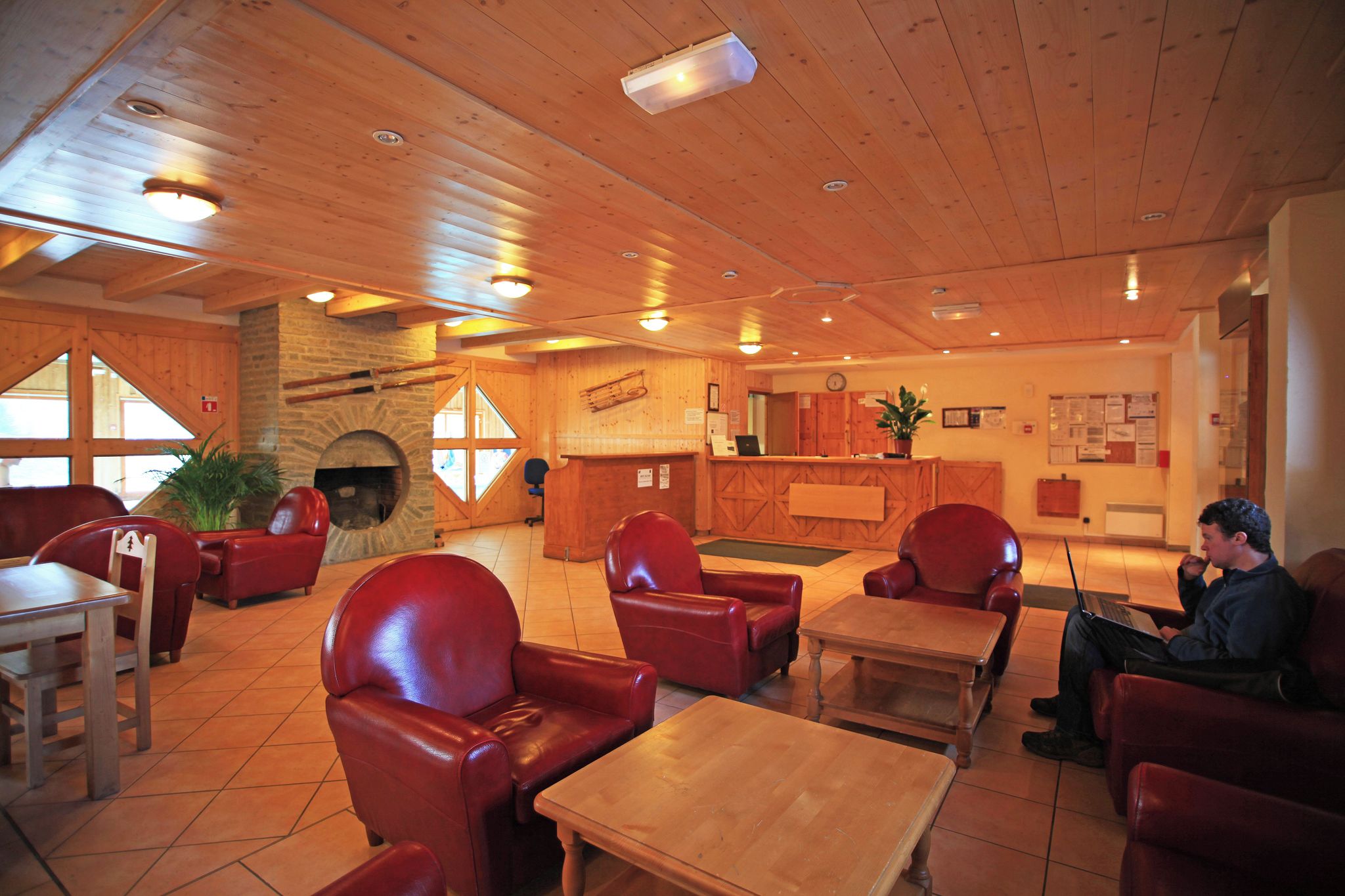 A cozy interior with wooden ceilings, red leather chairs, a fireplace, a reception desk, and a laptop on a table.