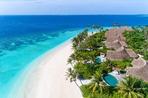 Vista aérea de un complejo turístico en la playa con villas de techo de paja, piscina y mar turquesa transparente.
