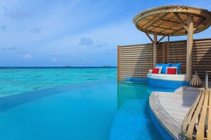 An infinity pool with a wooden deck and a lounge area with a thatched roof and colorful cushions.