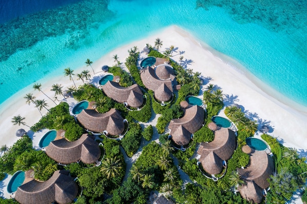 Vista aérea de un resort con bungalows de techo de paja, piscinas y una playa.