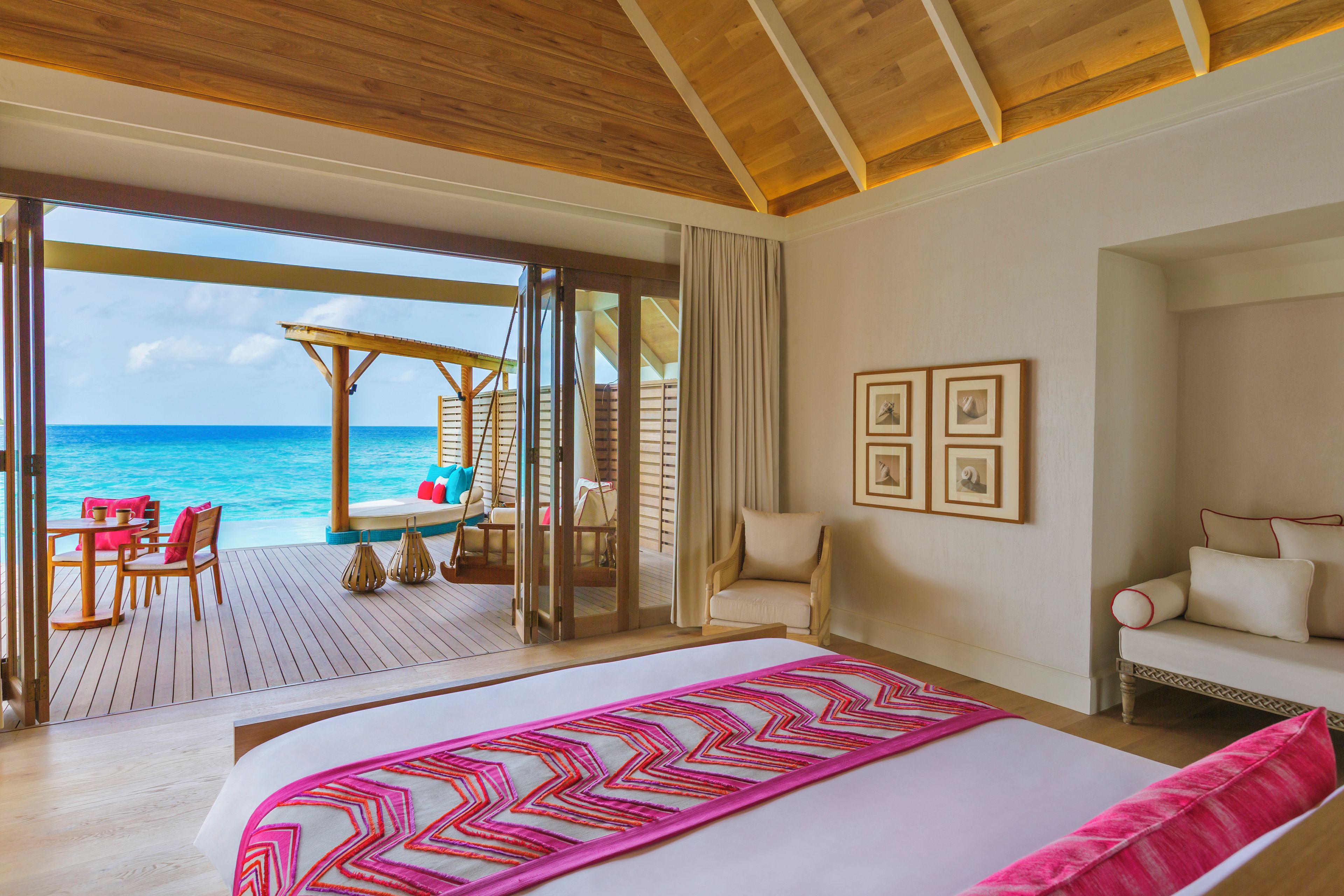A hotel room with a large bed, a view of the ocean, a balcony with a table and chairs, and a wooden ceiling.