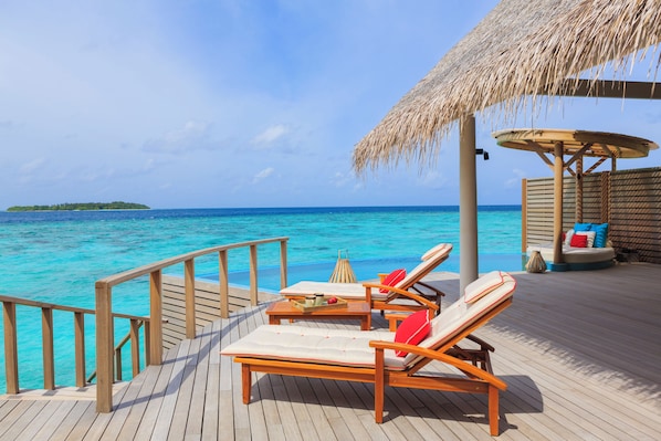 A wooden deck with lounge chairs and a thatched umbrella, overlooking a clear blue sea.