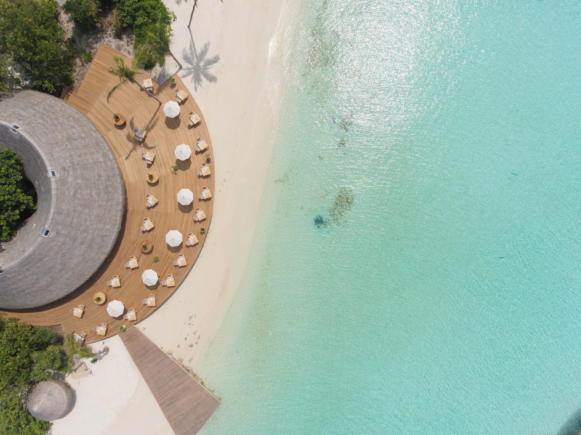 An aerial view of a beach resort with a circular wooden deck, lounge chairs, and umbrellas.