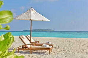 A beach with two wooden lounge chairs under a white umbrella, a sandy beach, and a distant island. - Milaidhoo Island Maldives (Milaidhoo Island)