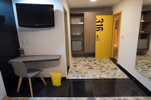 Habitación de hotel con televisión de pantalla plana, un escritorio con una silla, un cubo amarillo, piso con diseño de piedra y una puerta de madera con el número 316.