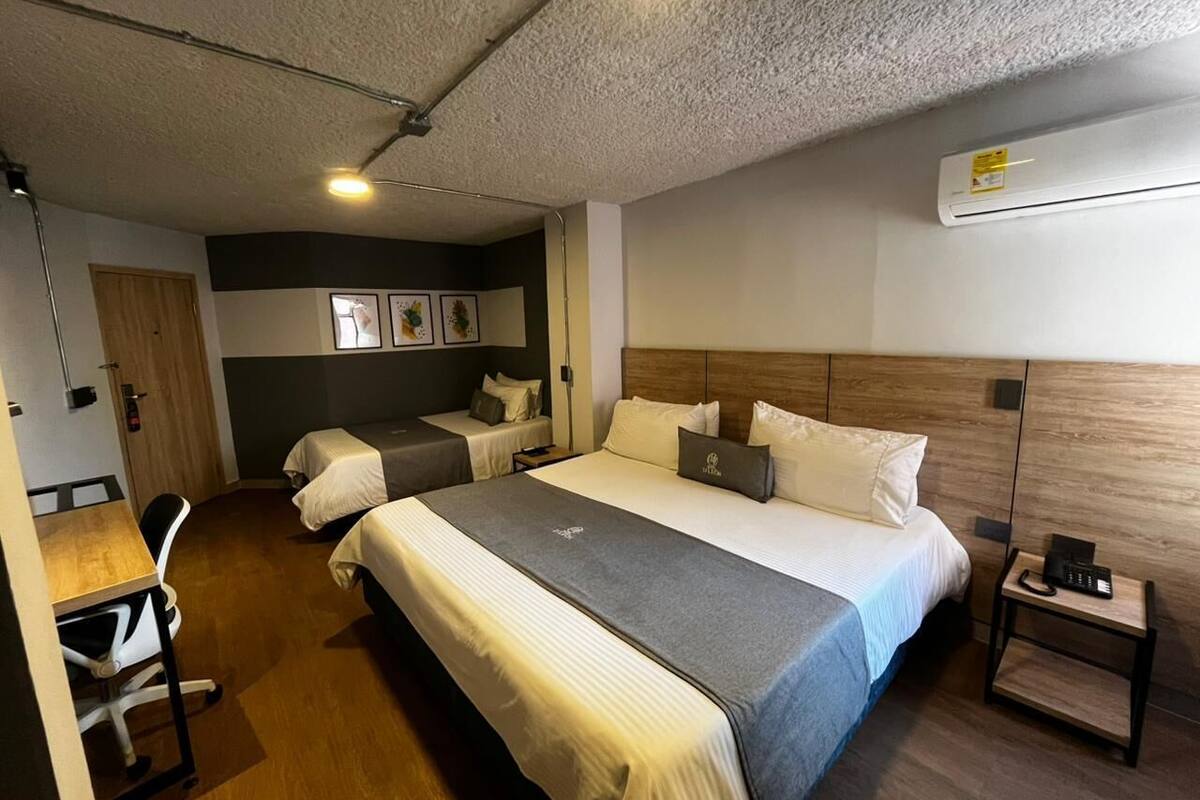Quarto de hotel com uma cama grande, uma escrivaninha, uma cadeira, mesa de cabeceira com telefone e ar-condicionado fixado na parede.