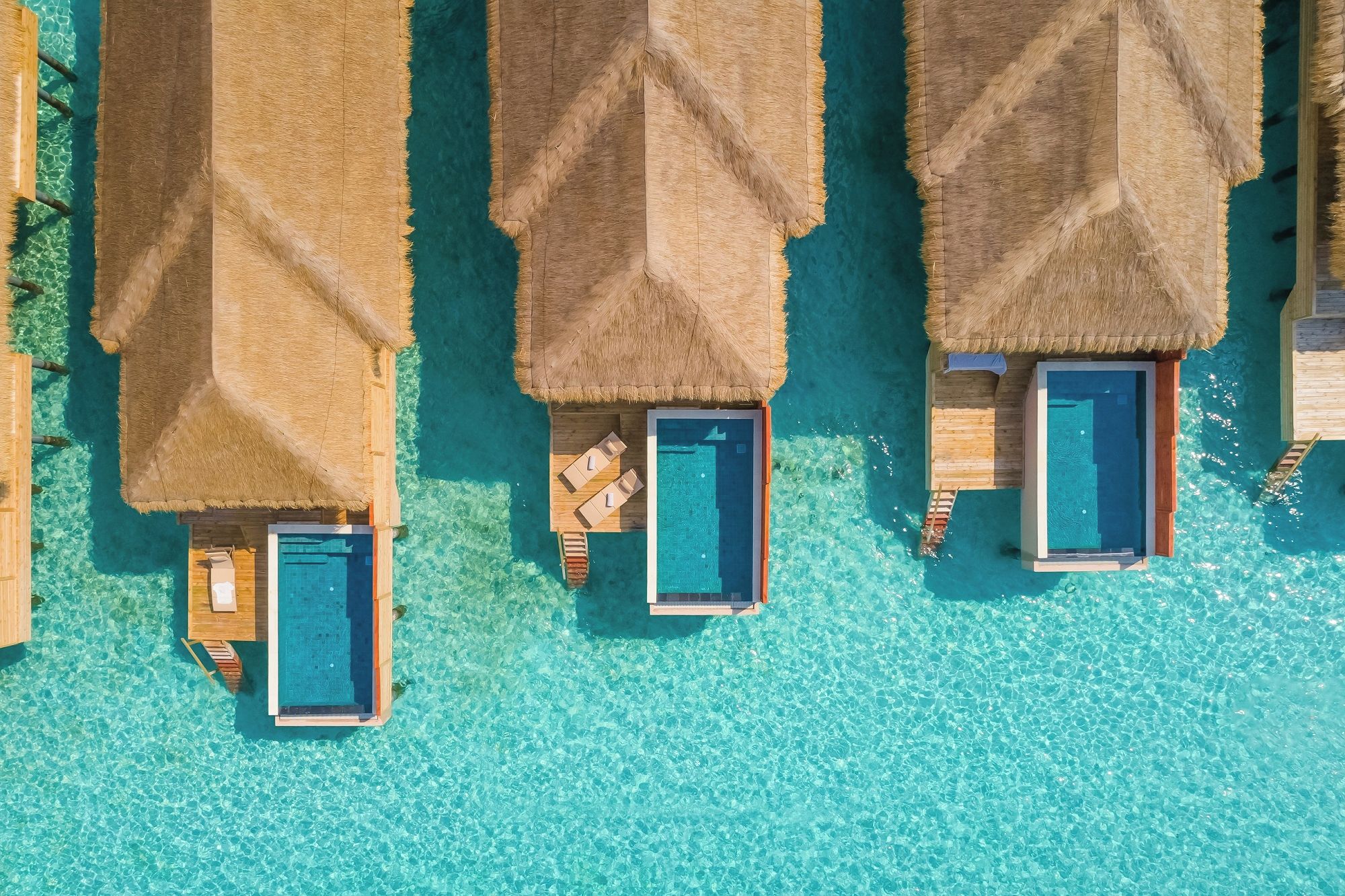 An aerial view of overwater bungalows with private pools, clear turquoise water, and thatched roofs.