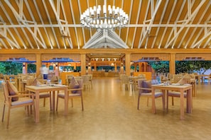 A spacious indoor dining area with wooden tables and chairs, a central chandelier, and a view of the outdoors through large windows.