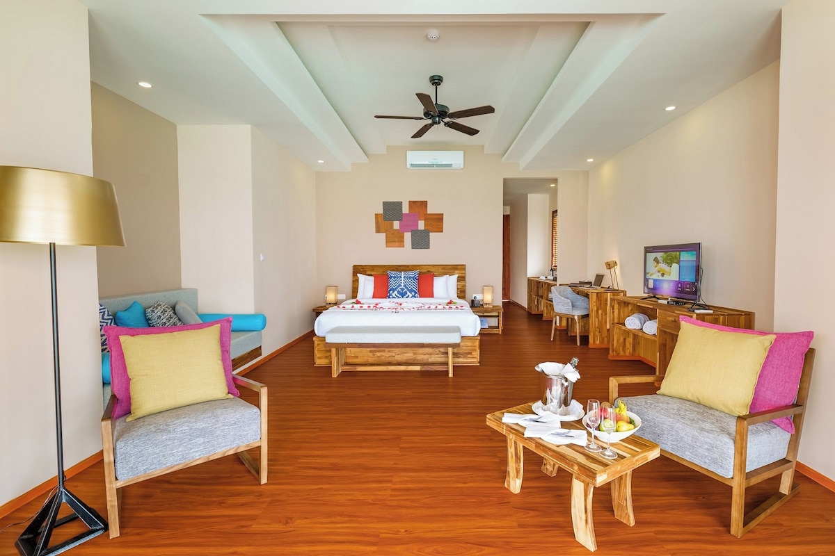 A hotel room with a bed, two armchairs, a desk, a television, and a ceiling fan.