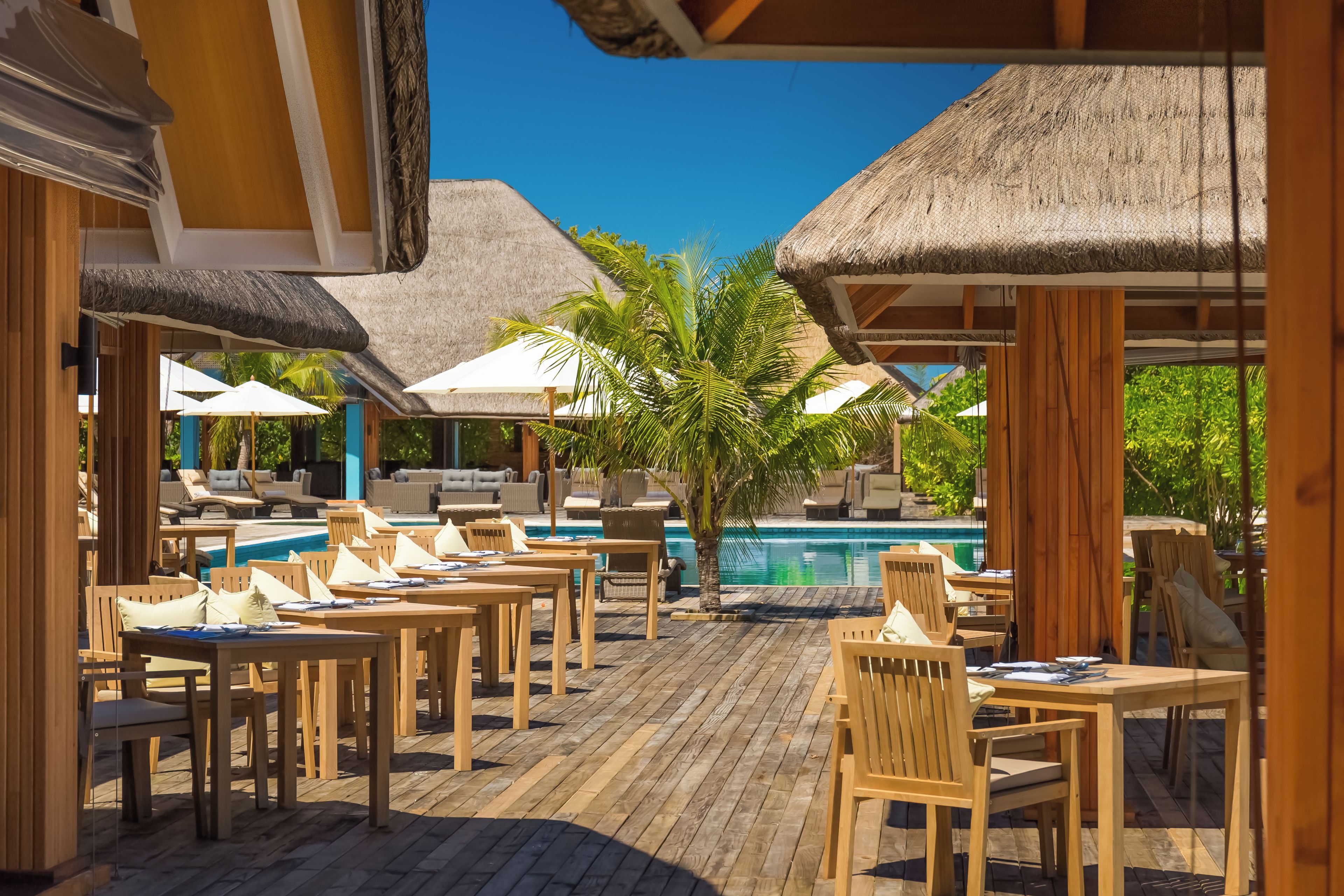 A poolside area with wooden tables and chairs, thatched umbrellas, and a clear blue sky.