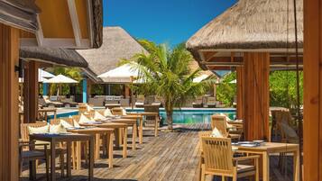 A poolside area with wooden tables and chairs, thatched umbrellas, and a clear blue sky.
