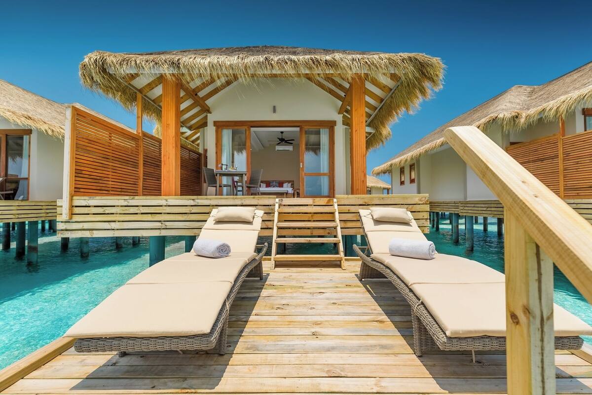 Una terraza de madera que lleva a una piscina con sillones reclinables, una estructura con techo de paja y una zona de comedor.