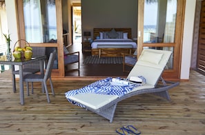 A wooden deck with a lounge chair, a wicker table with a basket of coconuts, and a bed with a patterned bedspread.