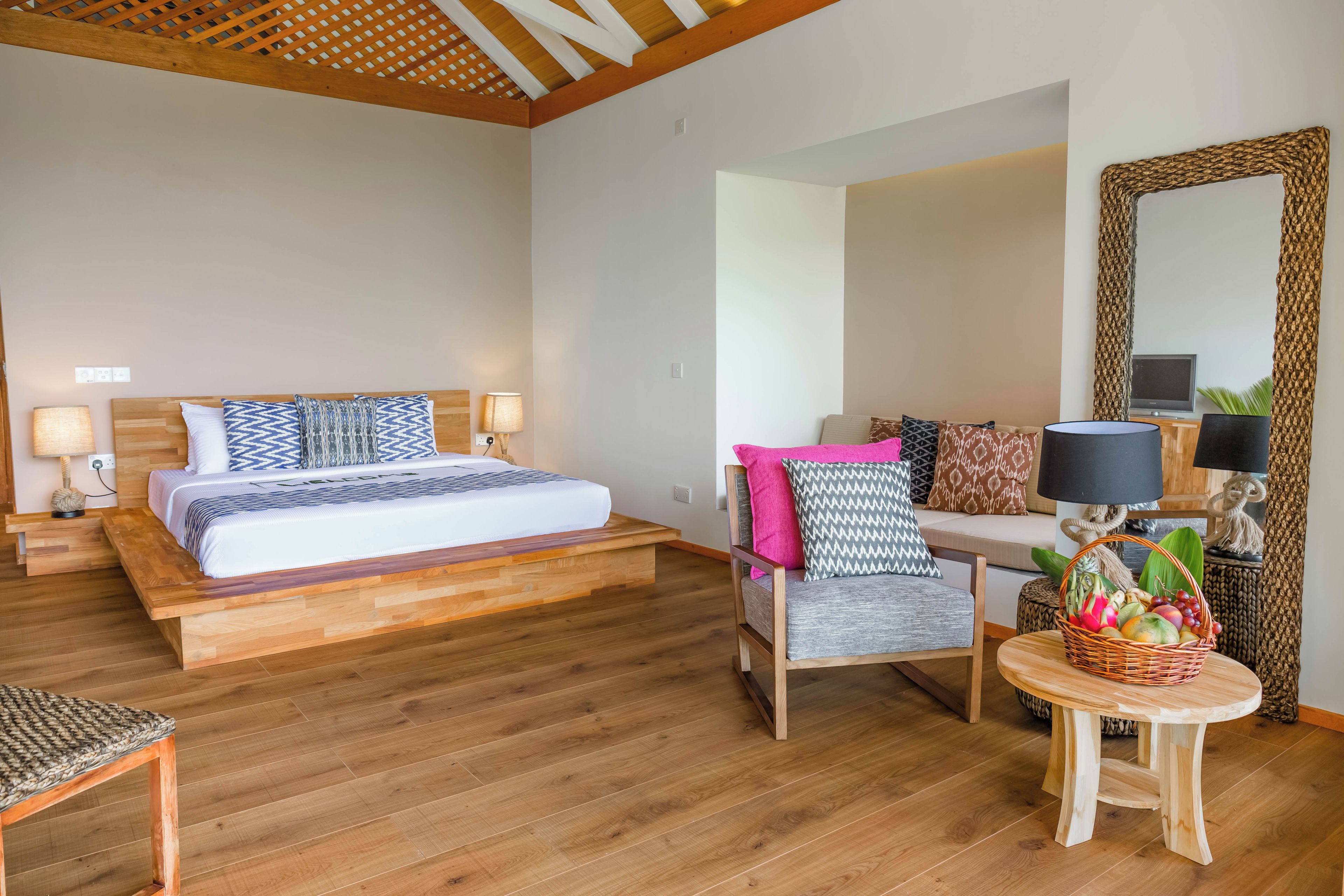 A hotel room with a wooden bed, a sofa, a small table with a fruit basket, and a large mirror.