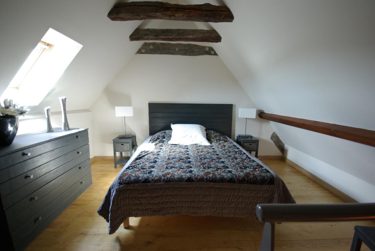 A bedroom with a sloped ceiling, a bed with a patterned bedspread, a dresser, two bedside tables with lamps, and a window.