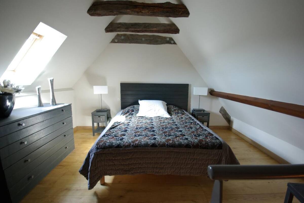 Un dormitorio con techo inclinado, una cama con colcha estampada, un aparador, dos mesitas de noche con lámparas y una ventana.