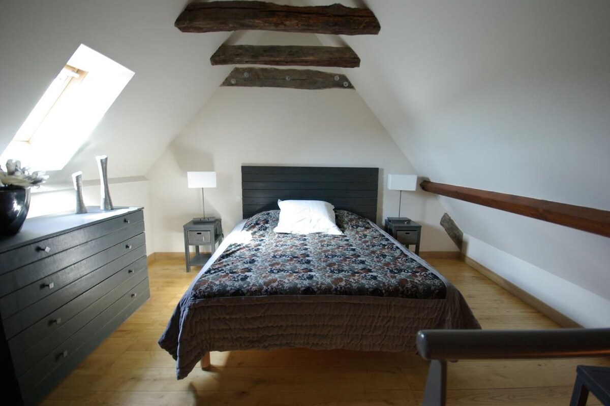 A bedroom with a sloped ceiling, a bed with a patterned bedspread, a dresser, two bedside tables with lamps, and a window.