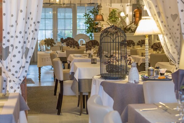 A dining area with tables set for service, a birdcage centerpiece, and a view of the dining room through a window.