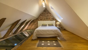 A bedroom with a sloped ceiling, a bed, a nightstand, a lamp, and a stone wall.