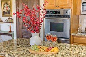 Una encimera de cocina con una bandeja de frutas, dos copas de vino rosado y un jarrón con flores rojas.