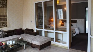 Ein Wohnzimmer mit Sofa, Hocker und Glastisch. Eine große Glasschiebetür führt in ein Schlafzimmer mit Bett und Nachttisch.