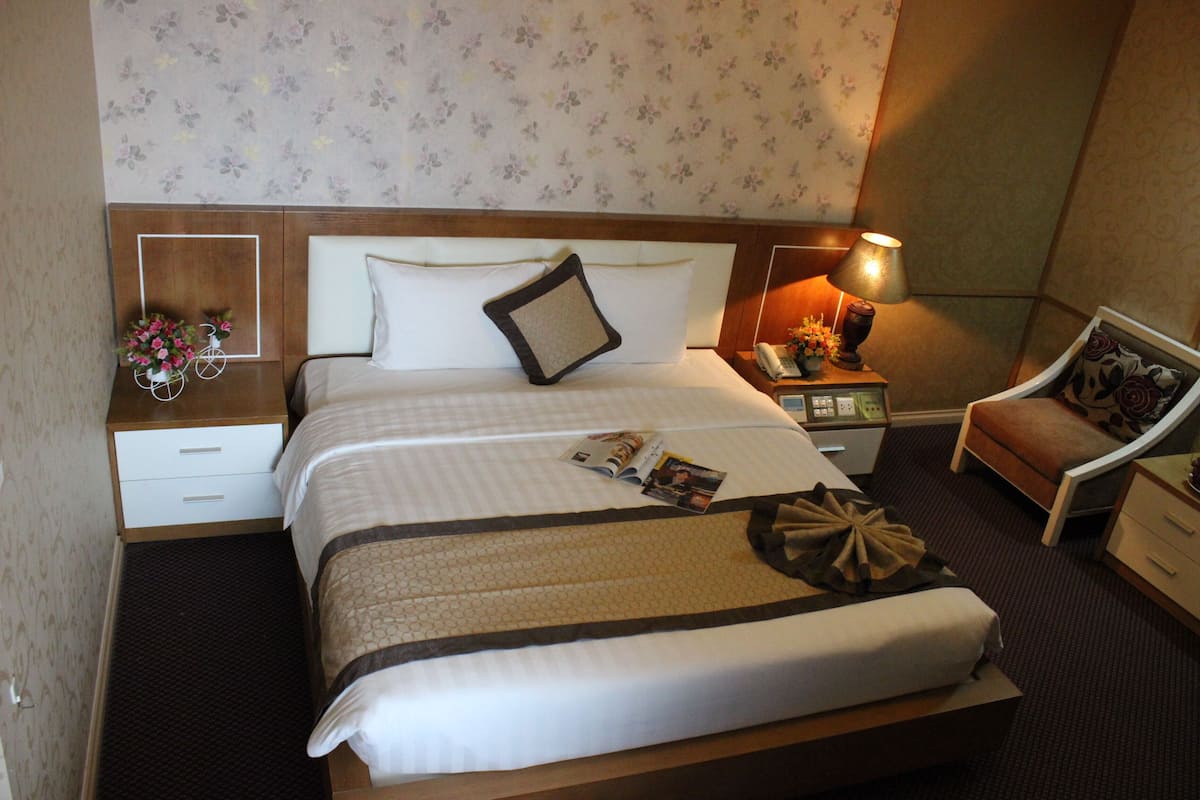 A hotel room with a large bed, a nightstand with a lamp, a chair, and a small table with a vase of flowers.