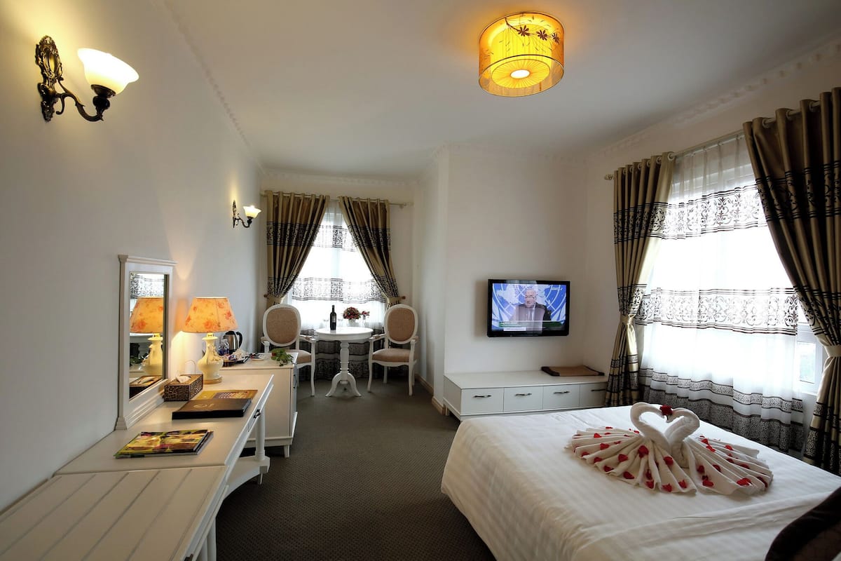 A hotel room with a bed, a desk with a lamp, a television, and a dining area with chairs and a table.
