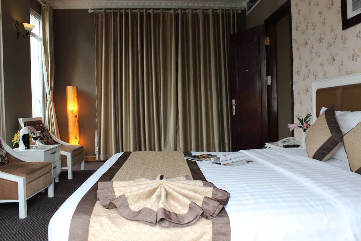 A hotel room with a large bed, a bedside table, a chair, a lamp, and a window with curtains.