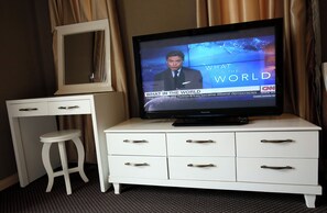 Habitación de hotel con una televisión mostrando un noticiero, un aparador blanco con cajones, un espejo y un taburete blanco pequeño.