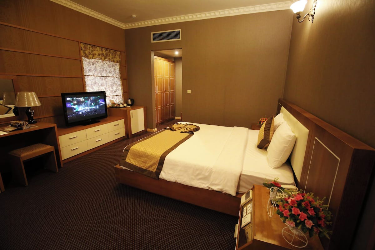 A hotel room with a bed, a television, a desk with a lamp, and a small bench.