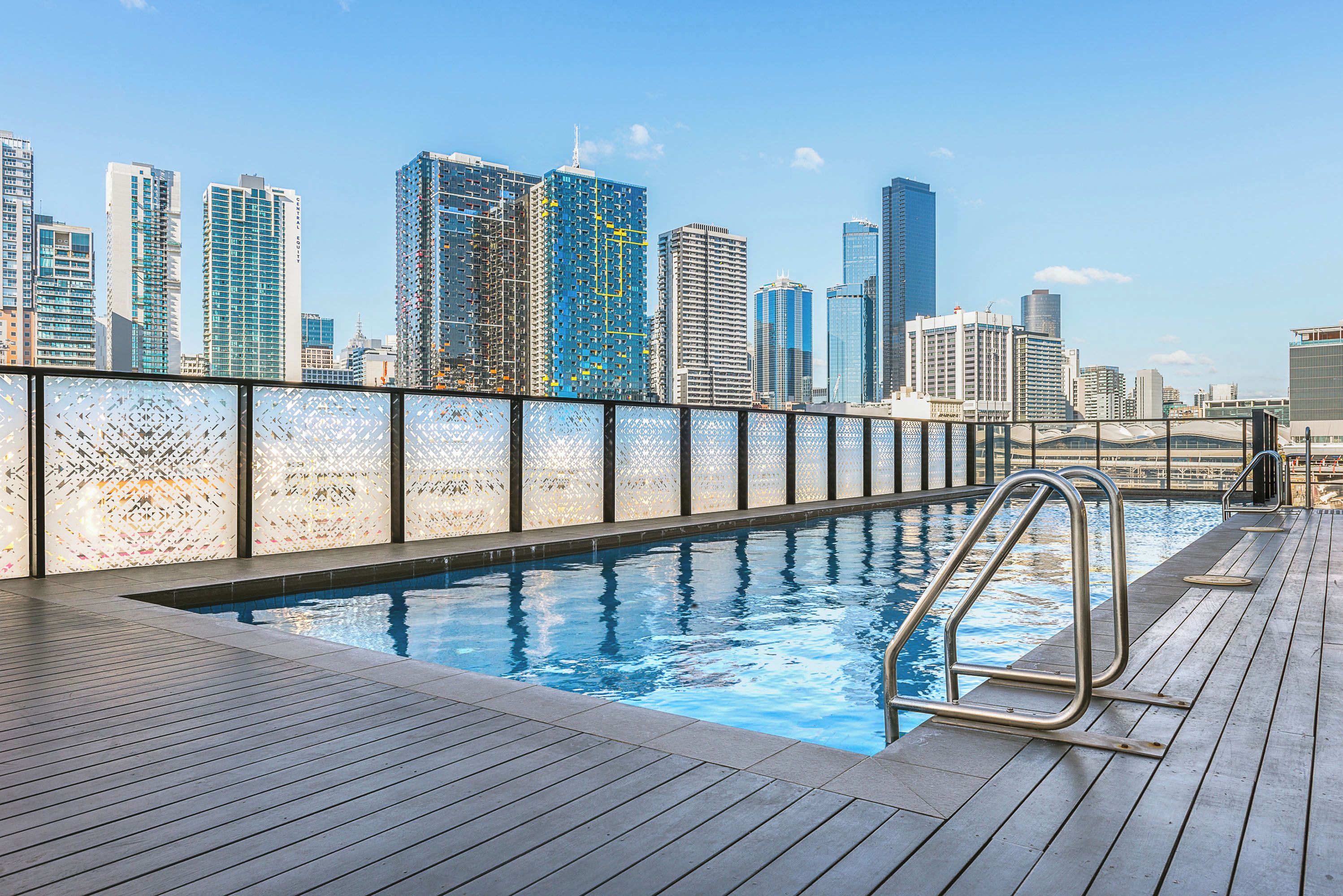 A rooftop pool with a city skyline view, a glass railing, and a wooden deck.
