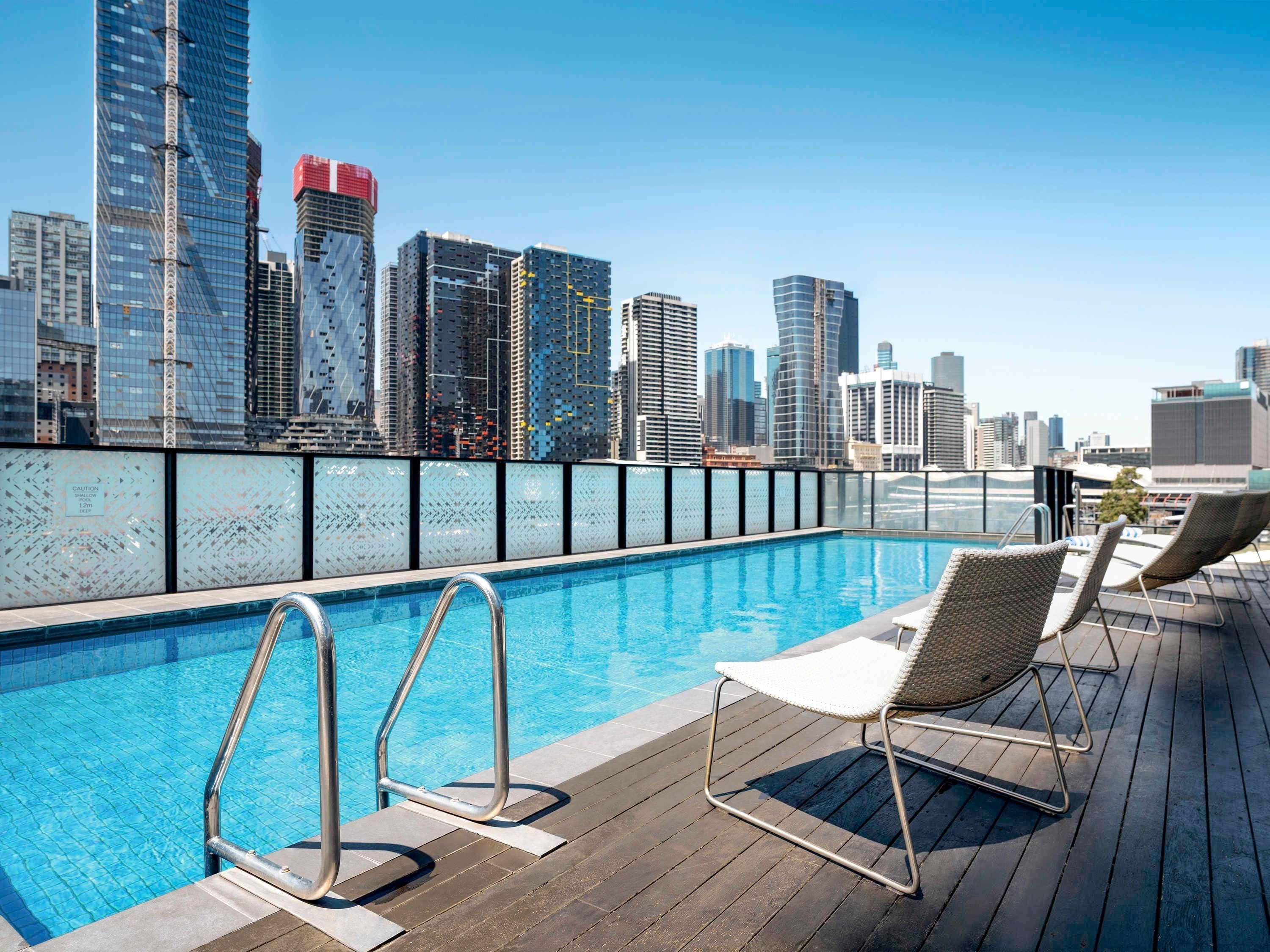 A rooftop pool with a city skyline view, metal pool ladders, and wicker lounge chairs.