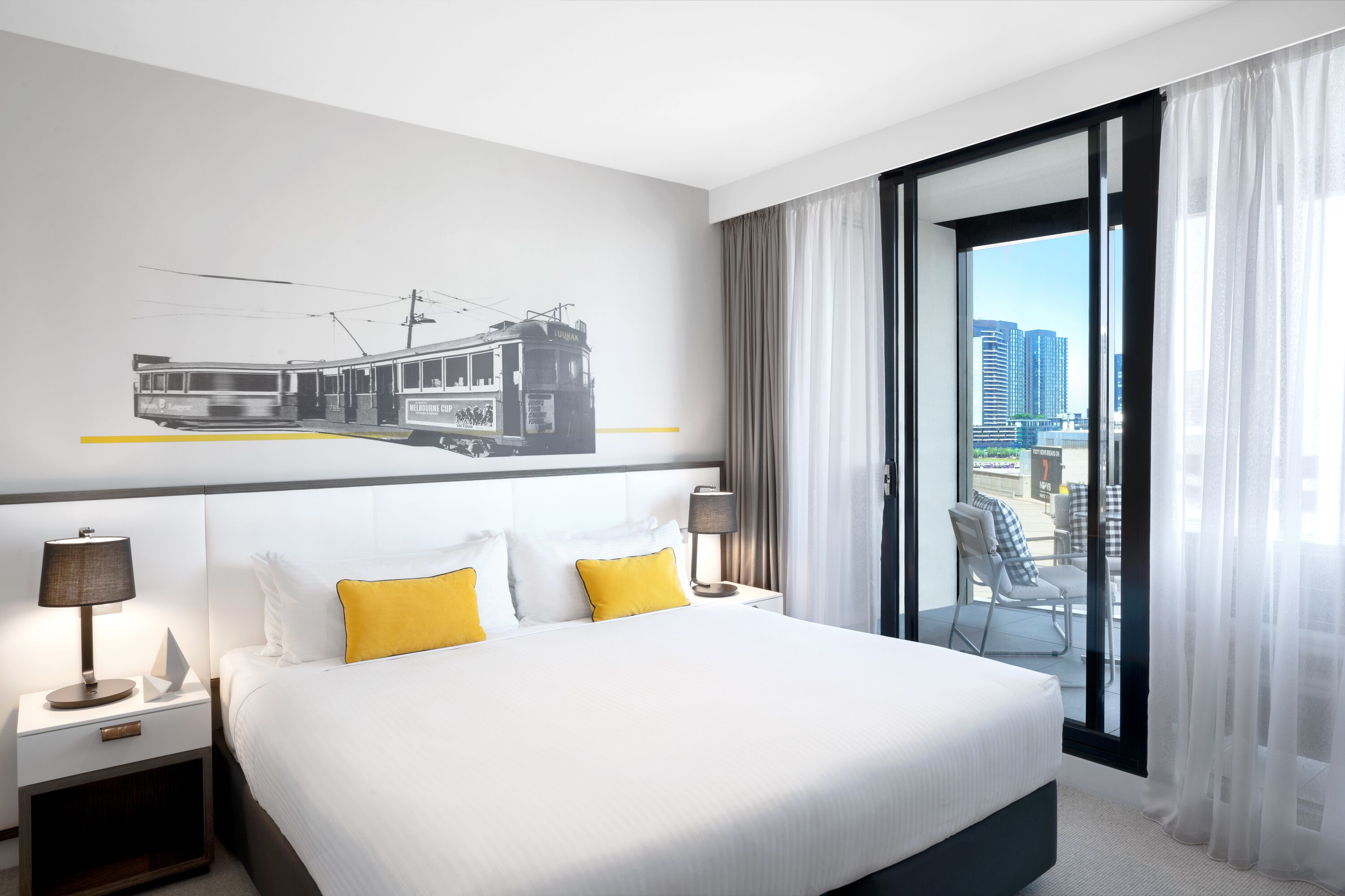 A hotel room with a bed, two bedside tables with lamps, a cityscape view through a large window, and a wall-mounted artwork of a tram.