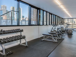 Ein Fitnessstudio mit Hanteln, Laufbändern und Trainingsgeräten, großen Fenstern und Blick auf die Stadt.