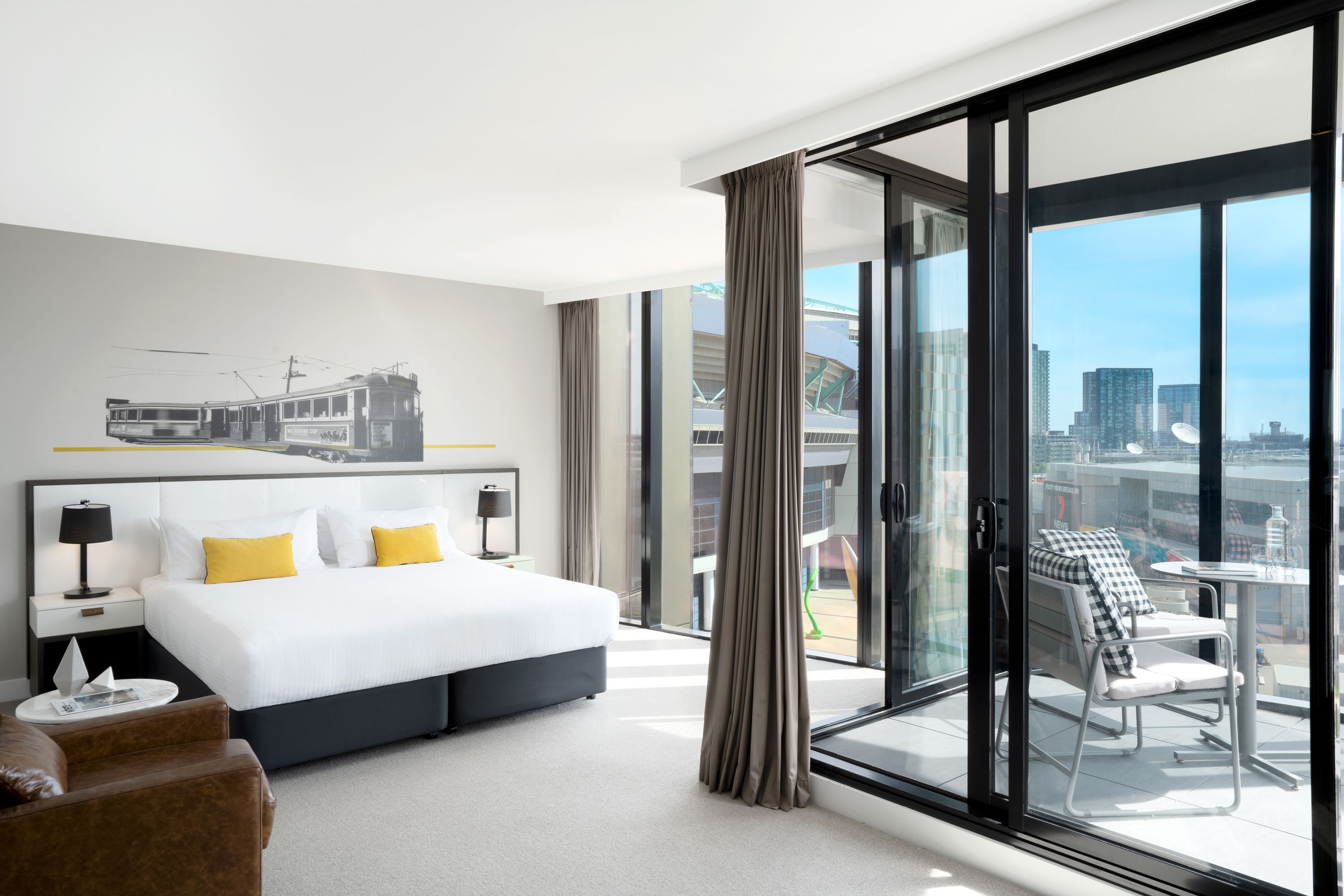 A modern hotel room with a large bed, a city view, a balcony with seating, and a window with curtains.