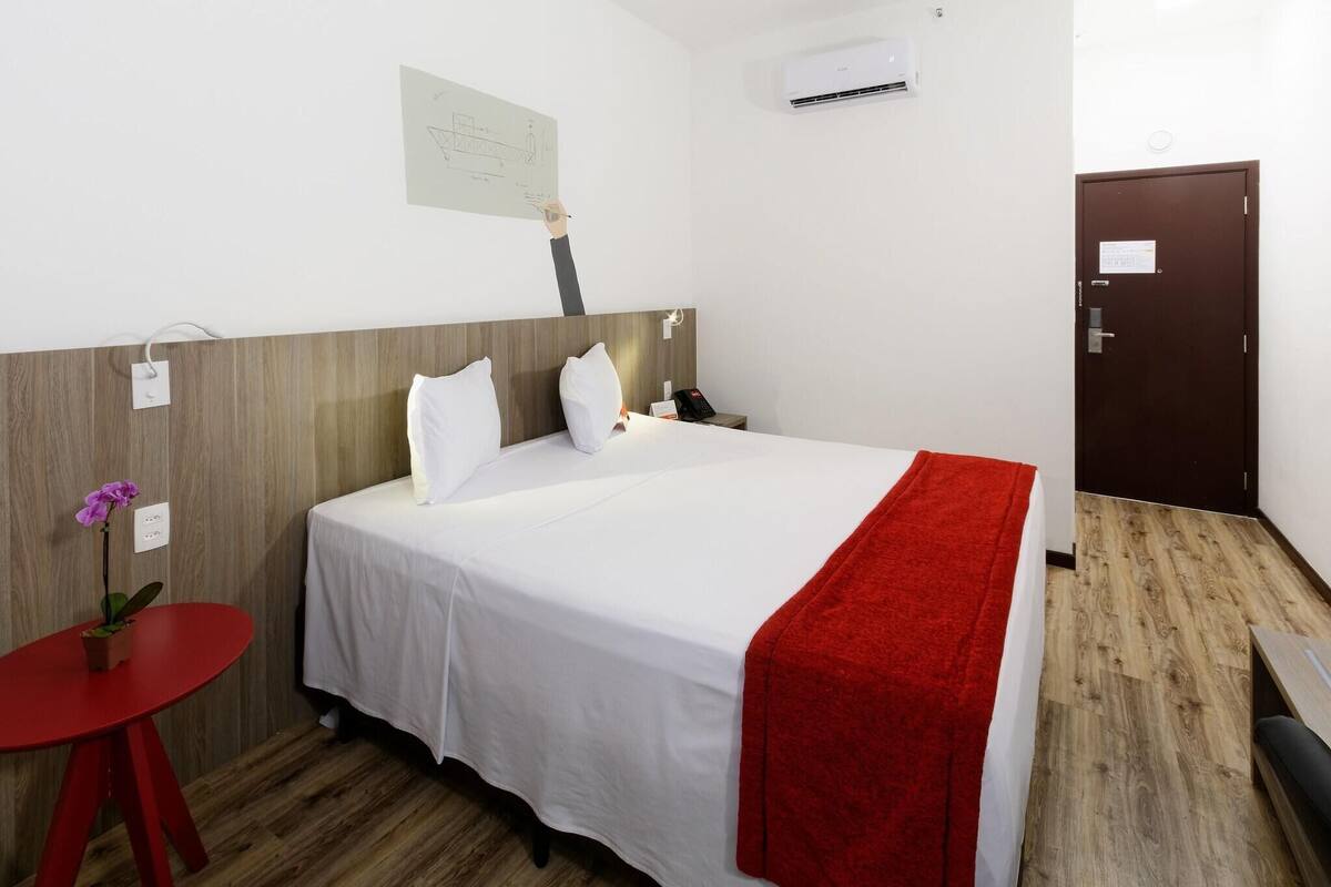 Quarto de hotel com uma cama, uma mesa vermelha com planta em vaso, ar condicionado na parede e uma porta.