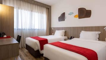 Quarto de hotel com duas camas, uma escrivaninha, uma cadeira e uma janela com cortinas.