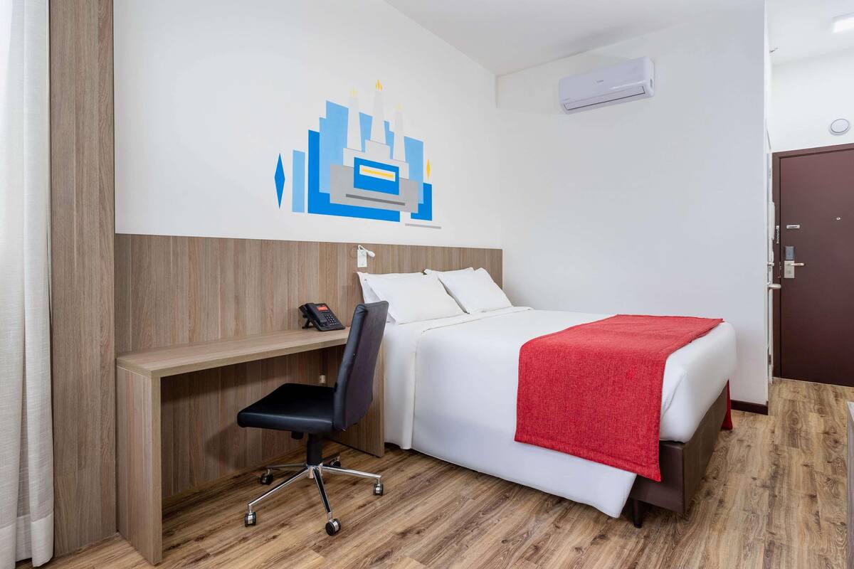 Quarto de hotel com cama, escrivaninha com cadeira, telefone e manta vermelha na cama.