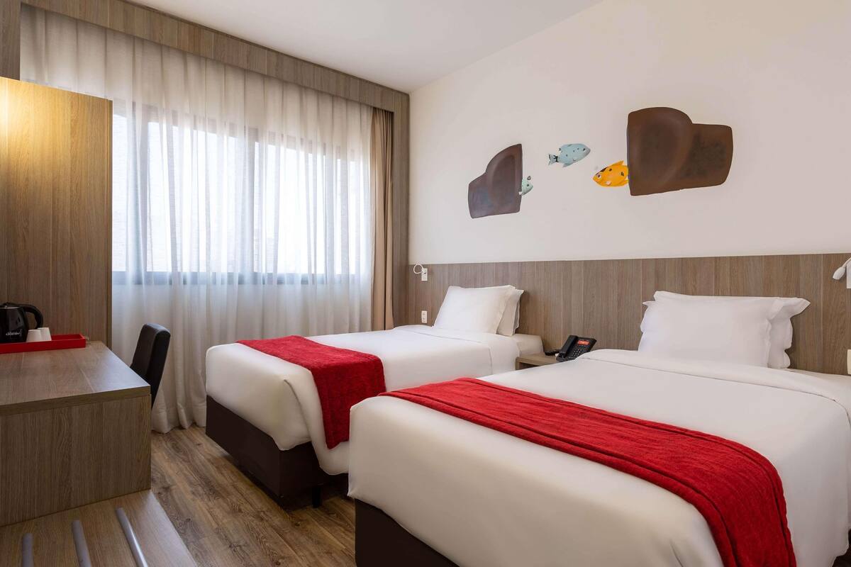 Quarto de hotel com duas camas, uma escrivaninha, uma cadeira e uma janela com cortinas.