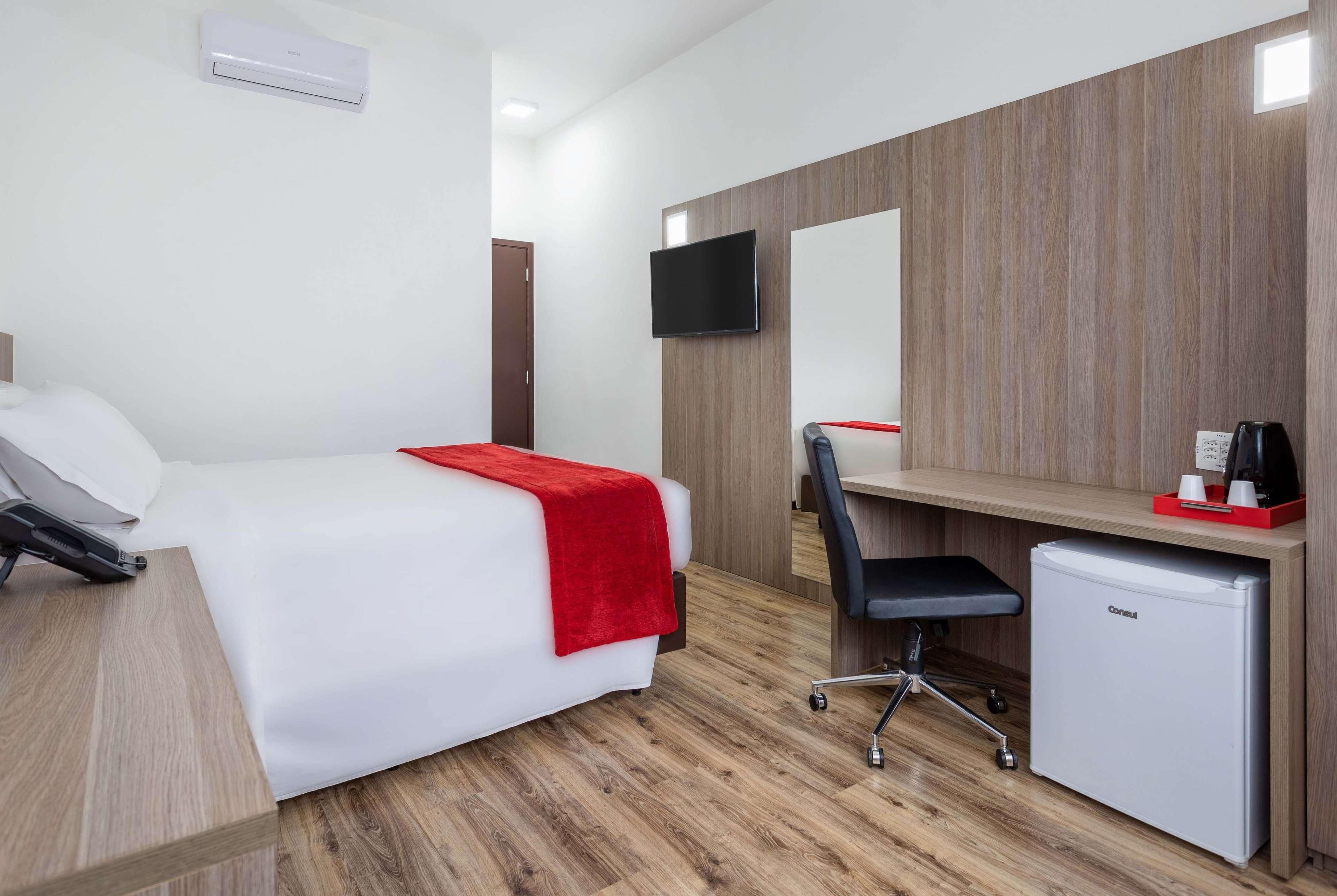 A hotel room with a bed, a desk, a chair, a small refrigerator, a TV, and an air conditioning unit.