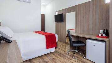A hotel room with a bed, a desk, a chair, a small refrigerator, a TV, and an air conditioning unit.